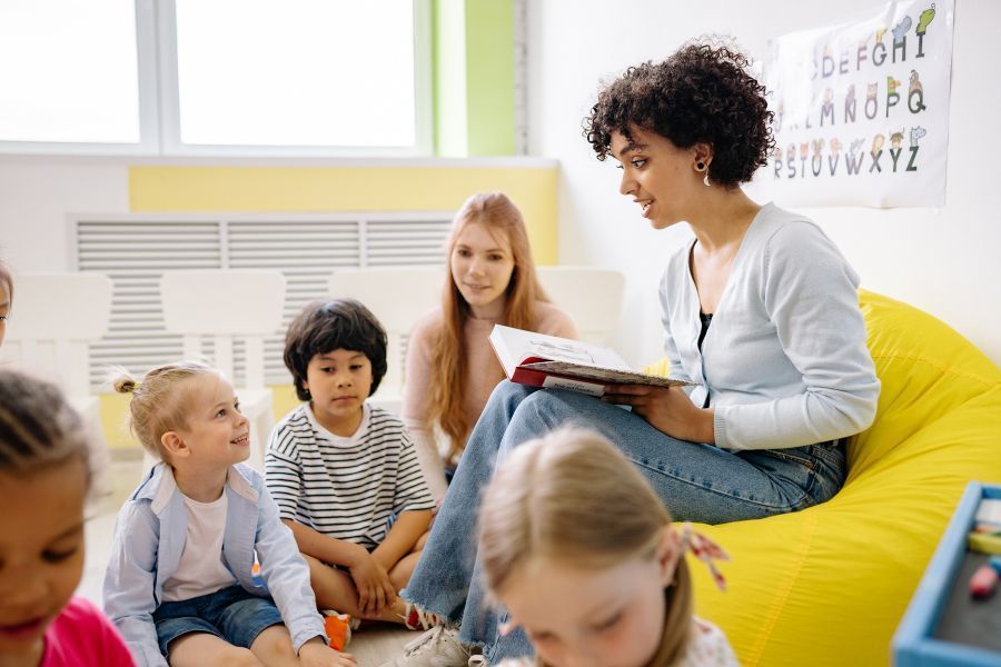 A teacher sits on a yellow beanbag chair, surrounded by young students seated on the floor, as she reads to them.