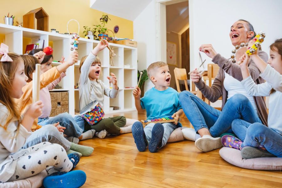 Preschool children gathered in a circle with their teacher, playing musical instruments in their classroom.